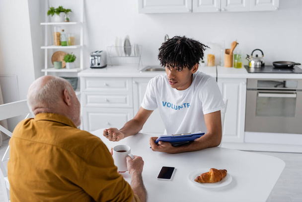 African american volunteer with clipboard talking to senior man with tea in kitchen  - Photo, image