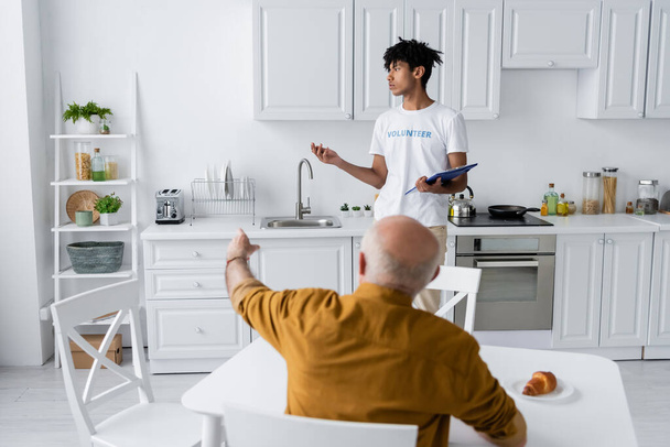 African american volunteer holding clipboard near blurred elderly man pointing with finger in kitchen  - 写真・画像