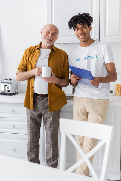 Smiling african american volunteer with clipboard and pensioner with cup looking at camera in kitchen  - Foto, Bild
