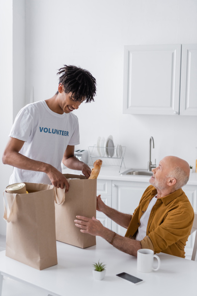 Smiling african american volunteer holding package with food near pensioner in kitchen  - Фото, изображение