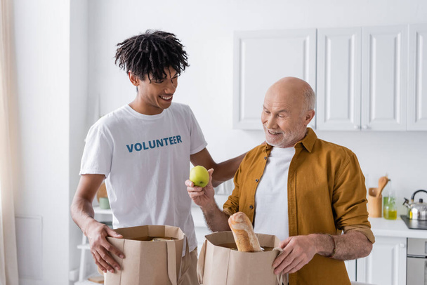 Усміхнений афроамериканський доброволець стоїть біля пенсіонера і пакує їжу на кухні
  - Фото, зображення