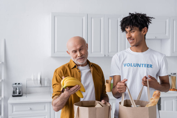 Smiling senior man holding bananas near bags and african american volunteer in kitchen  - Photo, Image