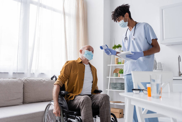 Старший мужчина в медицинской маске сидит в инвалидной коляске рядом с африканской американской медсестрой с пирометром и цифровым планшетом на кухне  - Фото, изображение