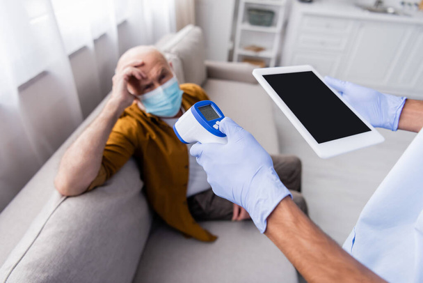 African american nurse holding digital tablet and pyrometer near blurred senior patient in medical mask at home  - 写真・画像