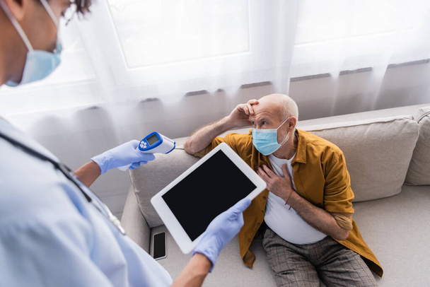 Пожилой человек в медицинской маске сидит рядом с размытой африканской американской медсестрой с пирометром и цифровым планшетом дома  - Фото, изображение