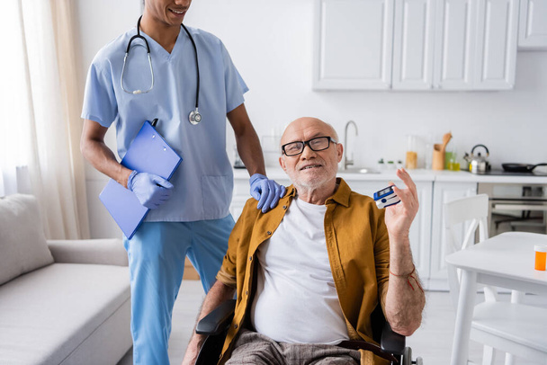 Smiling elderly man with pulse oximeter sitting in wheelchair near african american nurse with clipboard at home  - Foto, Imagen