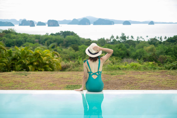 Šťastná žena v zelených plavkách plavat v luxusním bazénu hotelu na pozadí oceánu přírody, mladá žena s kloboukem těšit v tropickém letovisku. Relaxační, letní cestování, dovolená, dovolená koncept - Fotografie, Obrázek
