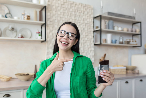 Young beautiful female nutritionist in glasses and green shirt advertises and recommends chocolate. He looks at the camera, points to the chocolate, poses, smiles. - Foto, immagini