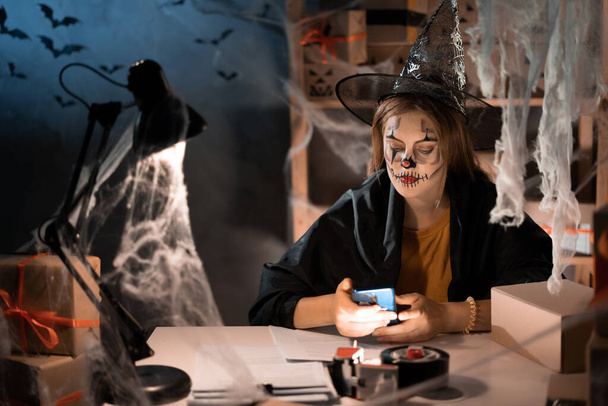Small business owner woman using mobile app on smartphone while checking parcel. Warehouse worker, seller in a witch costume holding a phone. Big sale discounts and promotions in honor of Halloween. - Foto, imagen