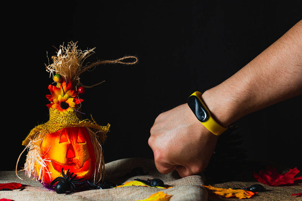 Arm with an activity bracelet with yellow wrist band next to a Halloween pumpkin adorned on sackcloth and surrounded by toy spiders and fallen leaves next to pine cones. - Photo, image