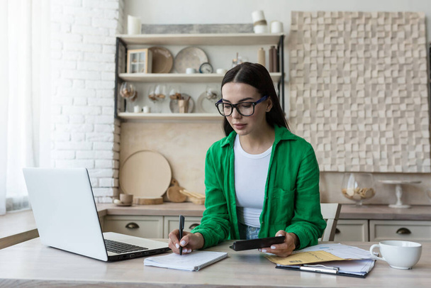 Home accounting.A young woman counts her income and expenses.checks the household budget. In glasses and a green shirt, sits at home with a laptop,a calculator and writes in a notebook,calculates. - Foto, Imagem