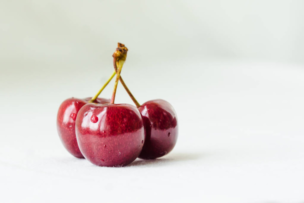 Three juicy berries of a red cherry with waterdrops on a white fabric background. Few red juicy cherries with stems. Delicious and ripe fruits - Photo, Image