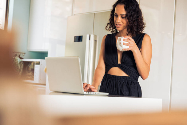 Smiling mid adult female freelancer holding coffee cup and working over laptop on kitchen island at home - Foto, Imagem