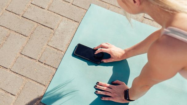 High angle cropped hands of caucasian young woman using mobile phone on yoga mat while exercising outdoors during sunny day - Fotó, kép