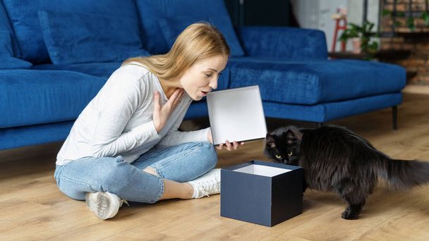 Ενθουσιασμένη γυναίκα που ανοίγει ένα κουτί δώρου, κάνοντας θαύματα και περίεργα πρόσωπα. Γυναίκα κάθεται στο πάτωμα στο άνετο σπίτι της, ελέγχοντας δώρο γενεθλίων μαζί με αστεία μαύρη γάτα της - Φωτογραφία, εικόνα