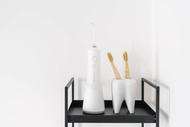 bamboo sensitive teeth brushes in holder, portable electric irrigator for mouth hygiene on metal black shelf in apartment bathroom with white wall, closeup - Foto, Imagem