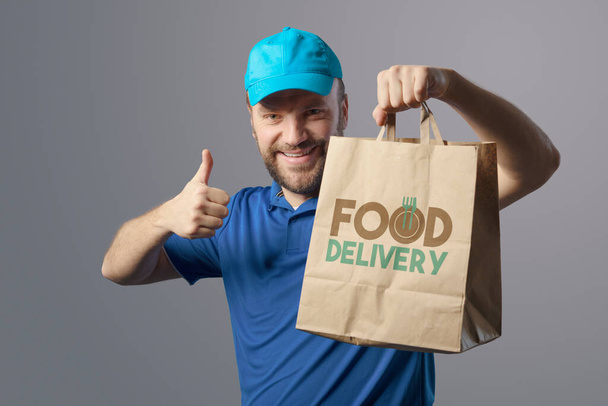 Smiling delivery guy holding a bag with fast food and giving a thumbs up - Фото, изображение