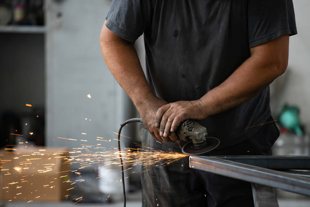 Handyman performing welding and grinding at his workplace in the workshop, while the sparks "fly" all around him. He is wearing a protective helmet and equipment. - Фото, изображение