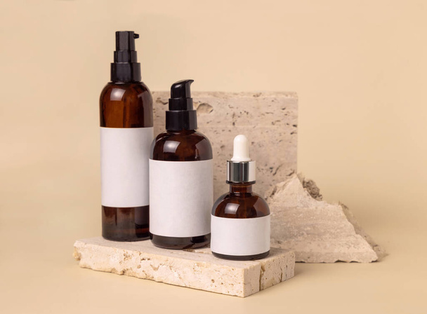 Brown glass one pump bottles on travertine stones on light beige close up, label mockup. Skincare beauty product package. Natural cosmetics, minimal compositio - Foto, Bild
