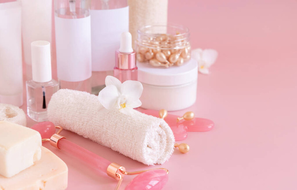 Frascos cosméticos naturais, acessórios de cuidados com a pele e massageadores de rosto de quartzo rosa com flor de orquídea branca em rosa claro de perto. Mockup de produtos de beleza. Mulher todos os dias enfrentam rotina de cuidados, tratamentos de spa - Foto, Imagem