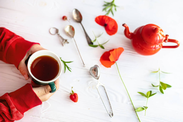 Hands holding a cup of coffee on background of white wooden table. Red teapot and fresh poppies during breakfast - Фото, зображення