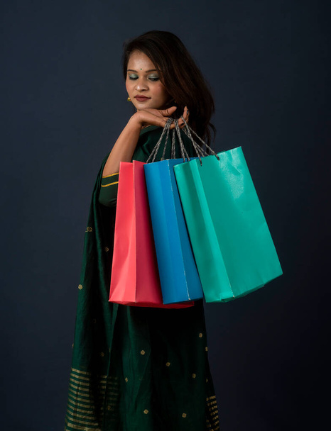 Beautiful Indian young girl or woman holding and posing with shopping bags on a dark background - 写真・画像