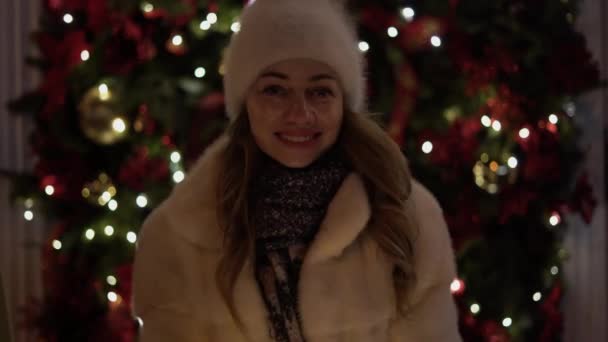 A beautiful young woman looks into the camera smiling and holds glowing garlands in her hands. Festive Christmas market on the background. Model looks at the camera, wears a knitted cap - Záběry, video