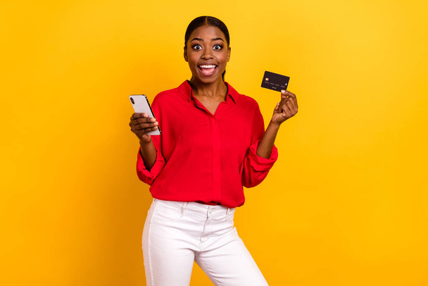 Photo of young excited girl shopping order bank card benefit cashback profit use mobile isolated over yellow color background. - Фото, изображение