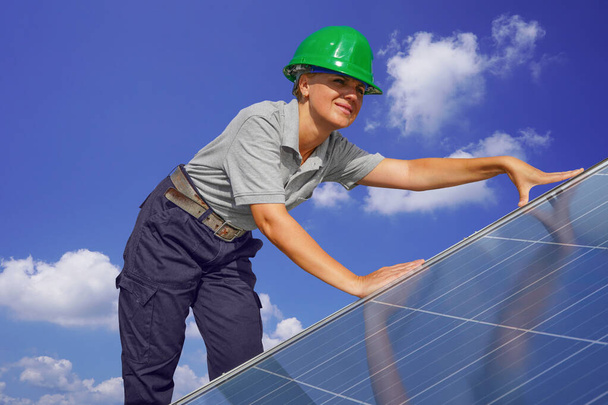 Woman engineer, electrician in green protective helmet against background of cloudy sky installing solar photovoltaic panel system. Professional electrician mounting solar panel. Alternative energy - Foto, Imagem