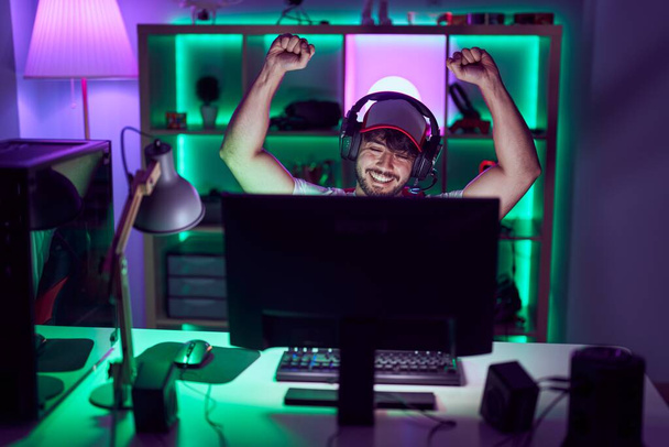Young hispanic man streamer using computer and headphones with winner expression at gamin room - Fotografie, Obrázek