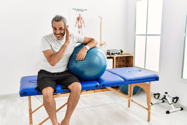 Middle age hispanic man at pain recovery clinic holding pilates ball hand on mouth telling secret rumor, whispering malicious talk conversation  - Photo, Image