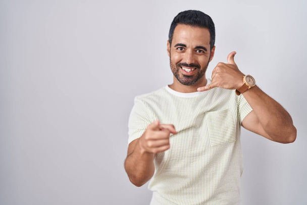 Hispanic man with beard standing over isolated background smiling doing talking on the telephone gesture and pointing to you. call me.  - Photo, Image
