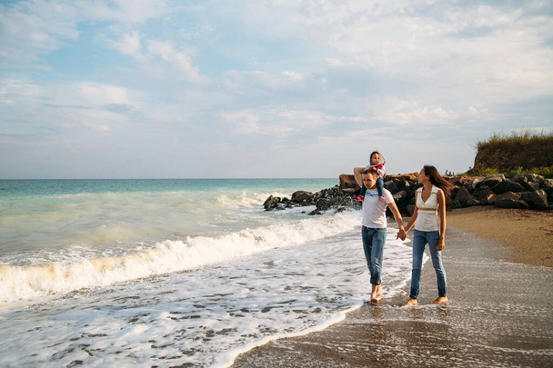 Счастливая молодая семья в белых футболках и синих джинсах с маленькой дочерью в синем платье, идущей вдоль моря. - Фото, изображение