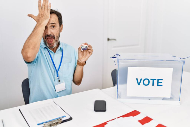 Middle age man with beard sitting by ballot holding i vote badge surprised with hand on head for mistake, remember error. forgot, bad memory concept.  - Foto, Imagem