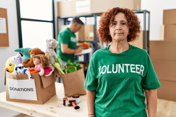 Middle age woman wearing volunteer t shirt at donations stand relaxed with serious expression on face. simple and natural looking at the camera.  - Zdjęcie, obraz