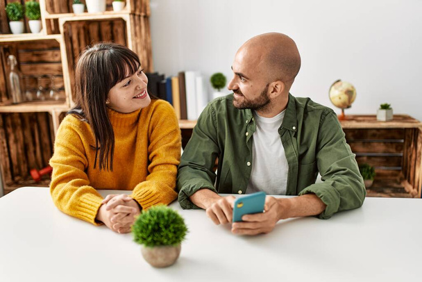 Молода пара іспаномовних посміхається щасливий, використовуючи смартфон, сидячи на столі вдома
. - Фото, зображення