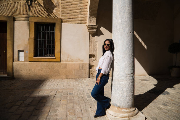 young, beautiful, brunette south american woman is travelling in europe and is leaning on a white marble column in a typical street of an old european city. Travel and holiday concept. - Fotografie, Obrázek