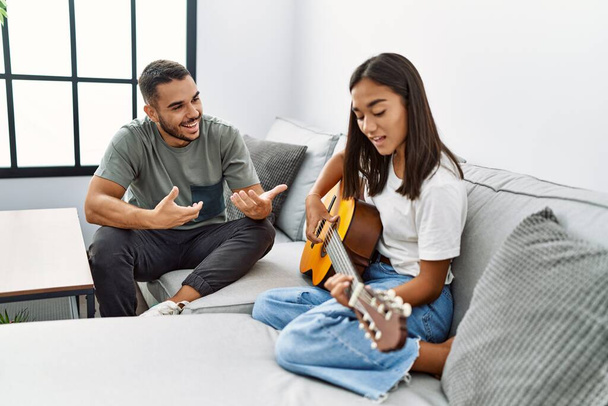 Joven pareja latina tocando la guitarra clásica sentada en el sofá en casa. - Foto, Imagen