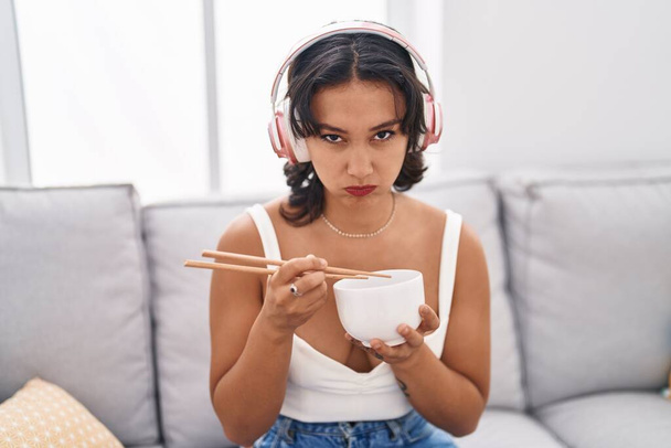 Молода іспаномовна жінка їсть азіатську їжу, використовуючи палички скептично і нервово, розчаровуючись через проблеми. негативна людина
.  - Фото, зображення