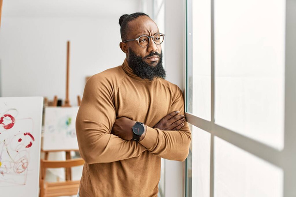 Νεαρός αφροαμερικανός καλλιτέχνης με σοβαρή έκφραση στέκεται με τα χέρια σταυρωμένα χειρονομία στο στούντιο τέχνης. - Φωτογραφία, εικόνα