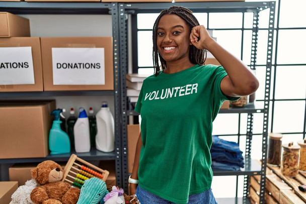 Fiatal afro-amerikai nő, aki önkéntes pólót visel adományoknál, mosolyogva áll fejre mutatva egy ujjal, nagyszerű ötlettel vagy gondolattal, jó memóriával.  - Fotó, kép