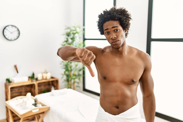 Jeune homme afro-américain torse nu portant serviette debout au centre de beauté regardant malheureux et en colère montrant rejet et négatif avec les pouces vers le bas geste. mauvaise expression.  - Photo, image