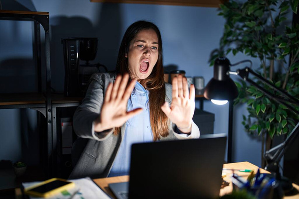 Fiatal barna nő dolgozik az irodában éjszaka fél, és retteg a félelem arckifejezéstől stop gesztus kézzel, kiabálva a sokk. pánikkoncepció.  - Fotó, kép