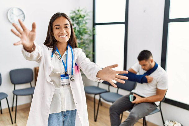 Young asian doctor woman at waiting room with a man with a broken arm looking at the camera smiling with open arms for hug. cheerful expression embracing happiness.  - Photo, Image