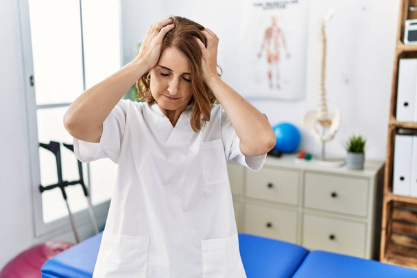 Középkorú fizioterapeuta nő dolgozik fájdalomcsillapító klinikán szenved fejfájás kétségbeesett és stresszes, mert a fájdalom és a migrén. kezeket a fejre..  - Fotó, kép