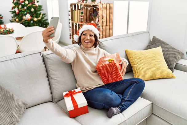 Hispanische Frau mittleren Alters macht Selfie mit dem Smartphone, wenn sie zu Hause auf dem Sofa am Weihnachtsbaum sitzt. - Foto, Bild