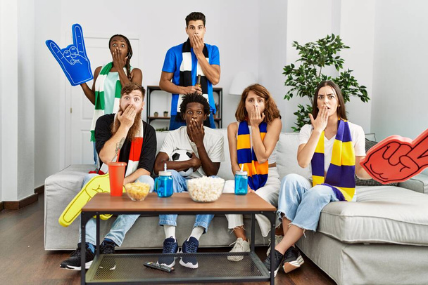 Group of friends supporting football team watching tv al home sitting on the sofa covering mouth with hand, shocked and afraid for mistake. surprised expression  - Photo, Image
