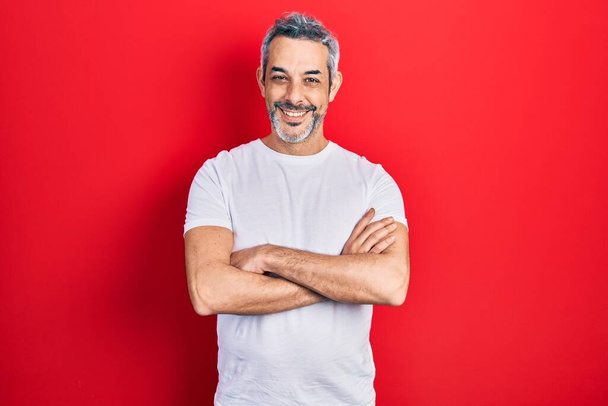 Ein gutaussehender Mann mittleren Alters mit grauen Haaren trägt ein lässiges weißes T-Shirt und lächelt mit verschränkten Armen in die Kamera. positive person.  - Foto, Bild