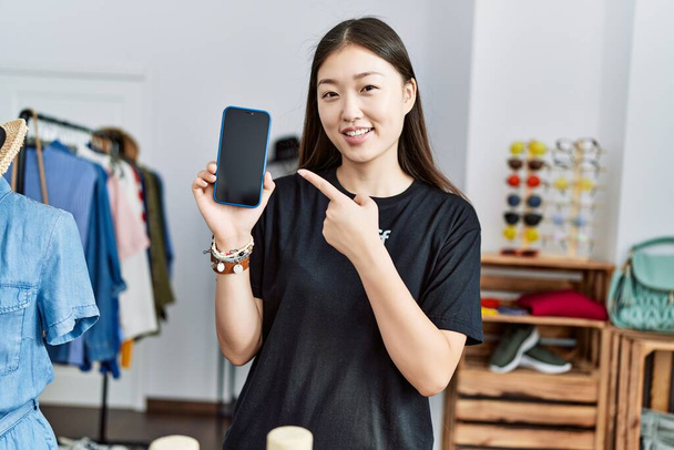 Jovem mulher asiática trabalhando como gerente no varejo boutique segurando smartphone sorrindo feliz apontando com mão e dedo  - Foto, Imagem