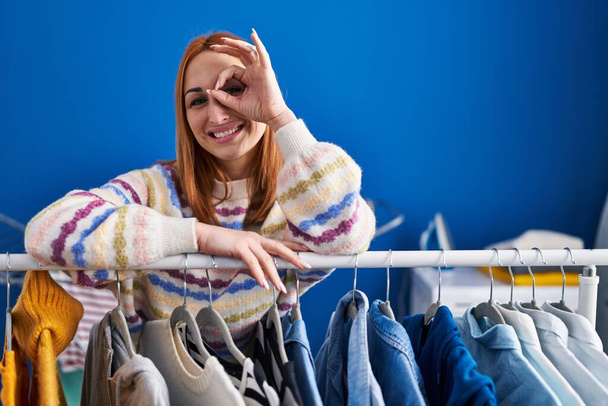 Mujer joven buscando ropa en perchero de ropa sonriendo feliz haciendo signo bien con la mano en el ojo mirando a través de los dedos  - Foto, Imagen